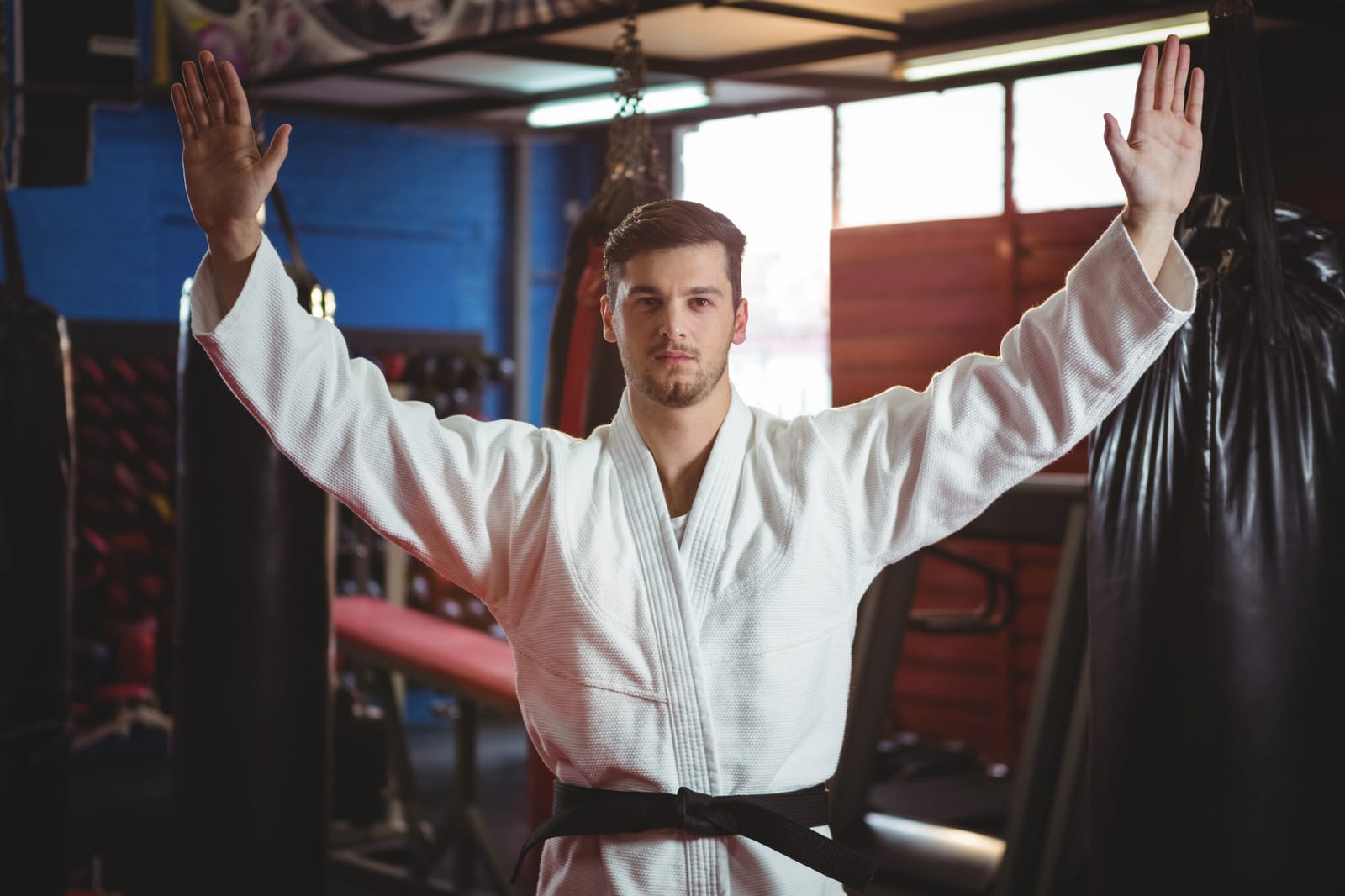 A student in a white gi and black belt practising a raised two-arm block among heavy bags.